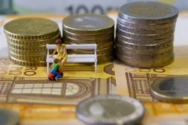 Auf einem Tisch liegt Geld und eine Mutter sitzt auf einer Bank. Vor ihr hockt ein kleines Kind