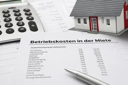 Dokument mit dem Titel „Betriebskosten in der Miete“ liegt auf einem Schreibtisch neben Taschenrechner, Stift und Modellhaus; aufgelistet sind typische Nebenkosten wie Wasser, Heizung und Müllabfuhr mit Eurobeträgen. 