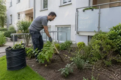 Mieter bei der Gartenarbeit vor einem Mehrfamilienhaus – er lockert die Erde im Beet und pflegt Pflanzen im gepflegten Außenbereich. 