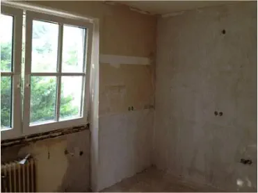 Ein Zimmer mit abgerissenen Tapeten, ein Fenster und eine Heizung, alles renovierungsbedürftig