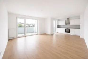 Heller, leerer Wohnraum mit sauberem Laminatboden, weißen Wänden und integrierter Küche – die Wohnung ist vollständig geräumt und besenrein übergeben, mit Zugang zu einem Balkon durch große Fensterfront. 